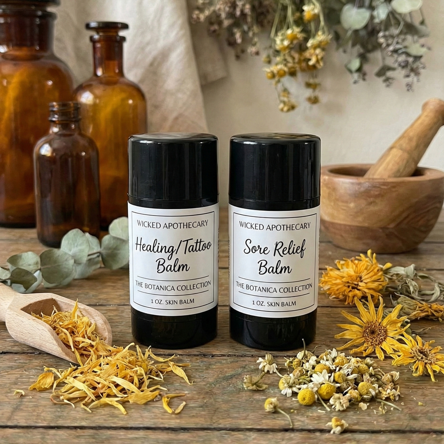 Two black balms labeled 'Healing Tattoo Balm' and 'Sore Relief Balm' on a wooden surface with dried herbs and bottles in the background.
