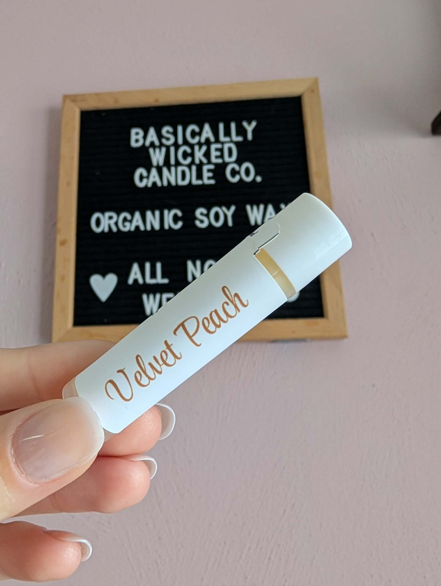Hand holding a small candle labeled 'Velvet Peach' in front of a sign reading 'Basically Wicked Candle Co.'