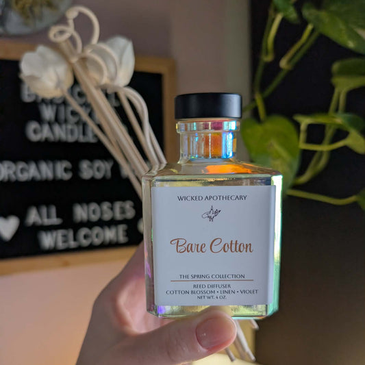 Bottle of 'Bare Cotton' by Wicked Apothecary held in front of a decorative background with plants and signs.