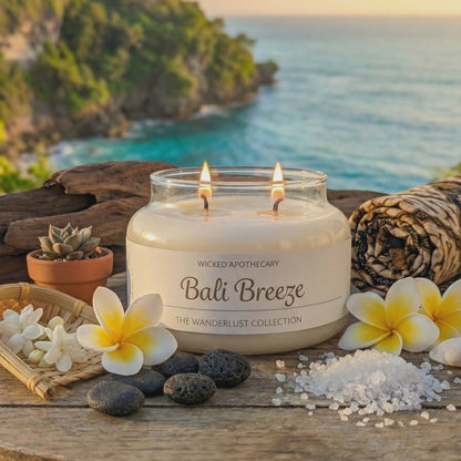 Candle labeled 'Bali Breeze' from Wicked Apothecary on a wooden table with flowers and stones, set against a scenic ocean backdrop.