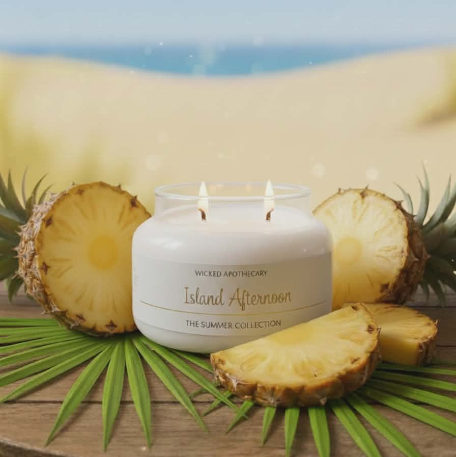 Candle labeled 'Island Afternoon' with pineapple slices and leaves on a wooden surface with a beach background.