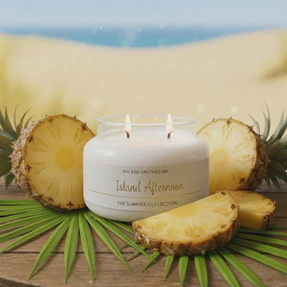 Candle labeled 'Island Afternoon' with pineapple slices and leaves on a wooden surface with a beach background.