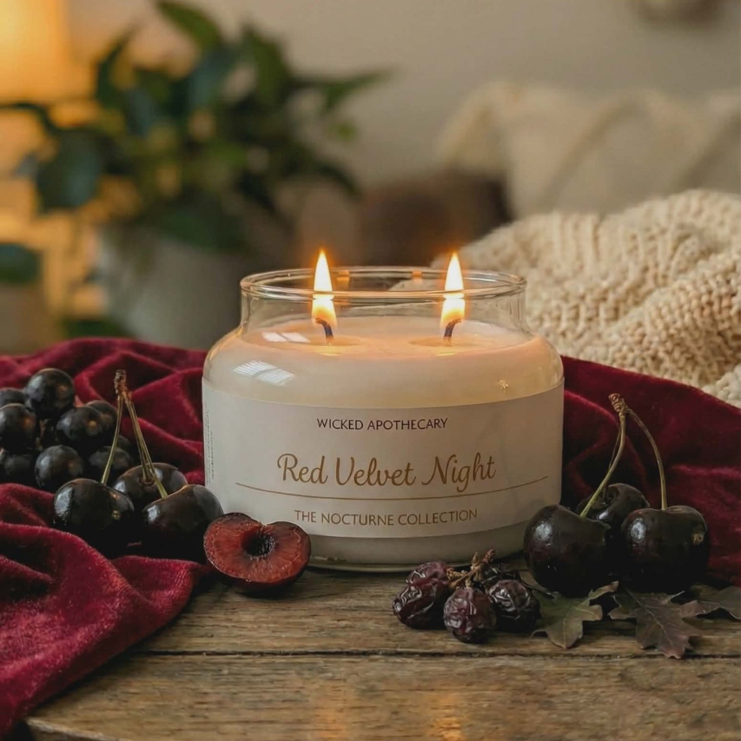 Candle in a glass jar labeled 'Red Velvet Night' on a wooden surface with cherries and a blurred background.