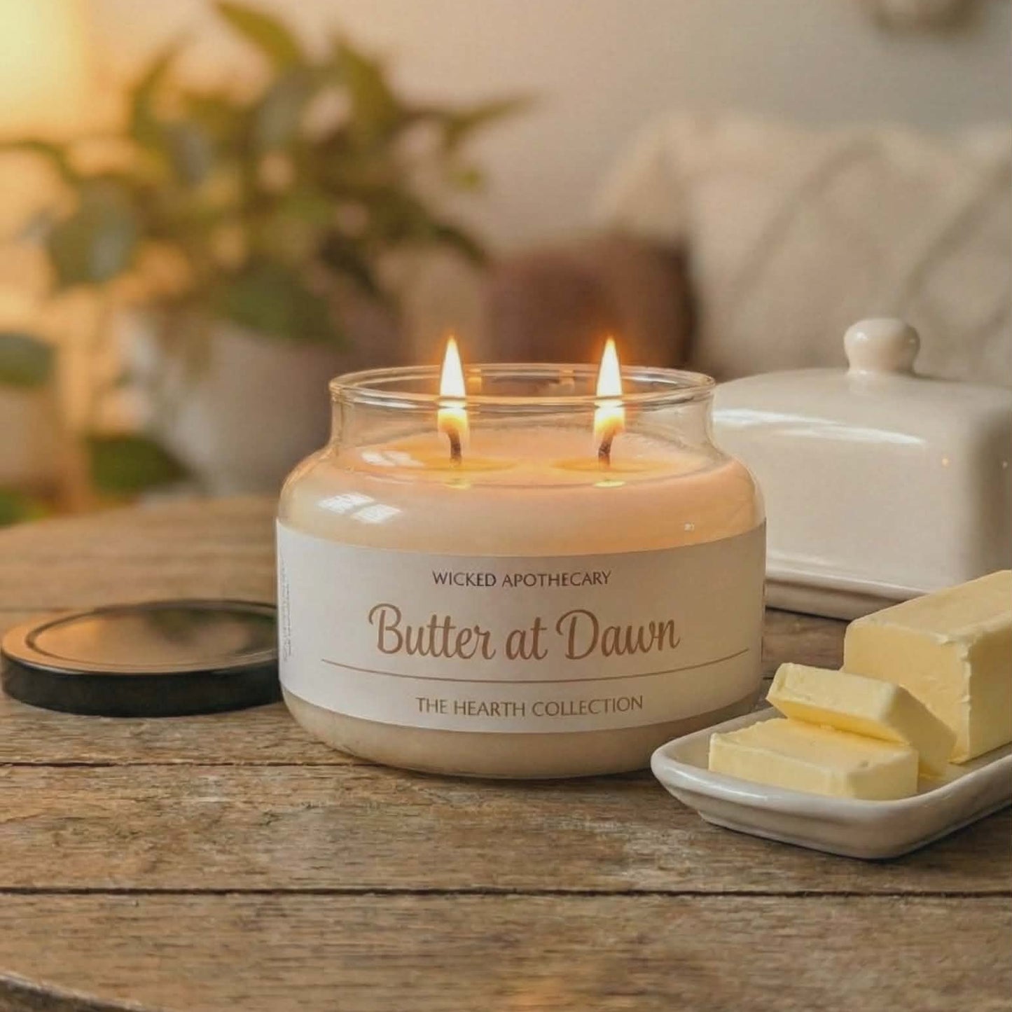 Two lit candles in a jar labeled 'Butter at Dawn' on a wooden table with a blurred background.