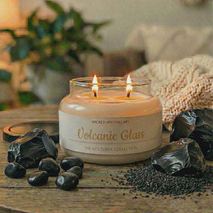 Candle in a glass jar labeled 'Volcanic Glass' on a wooden surface with decorative stones and plants.