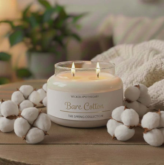 Candle labeled 'Bare Cotton' from Wicked Apothecary on a wooden surface with cotton flowers and blurred background