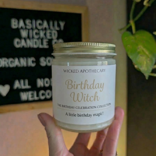 Candle jar labeled 'Birthday Witch' by Wicked Apothecary held in front of a store sign.