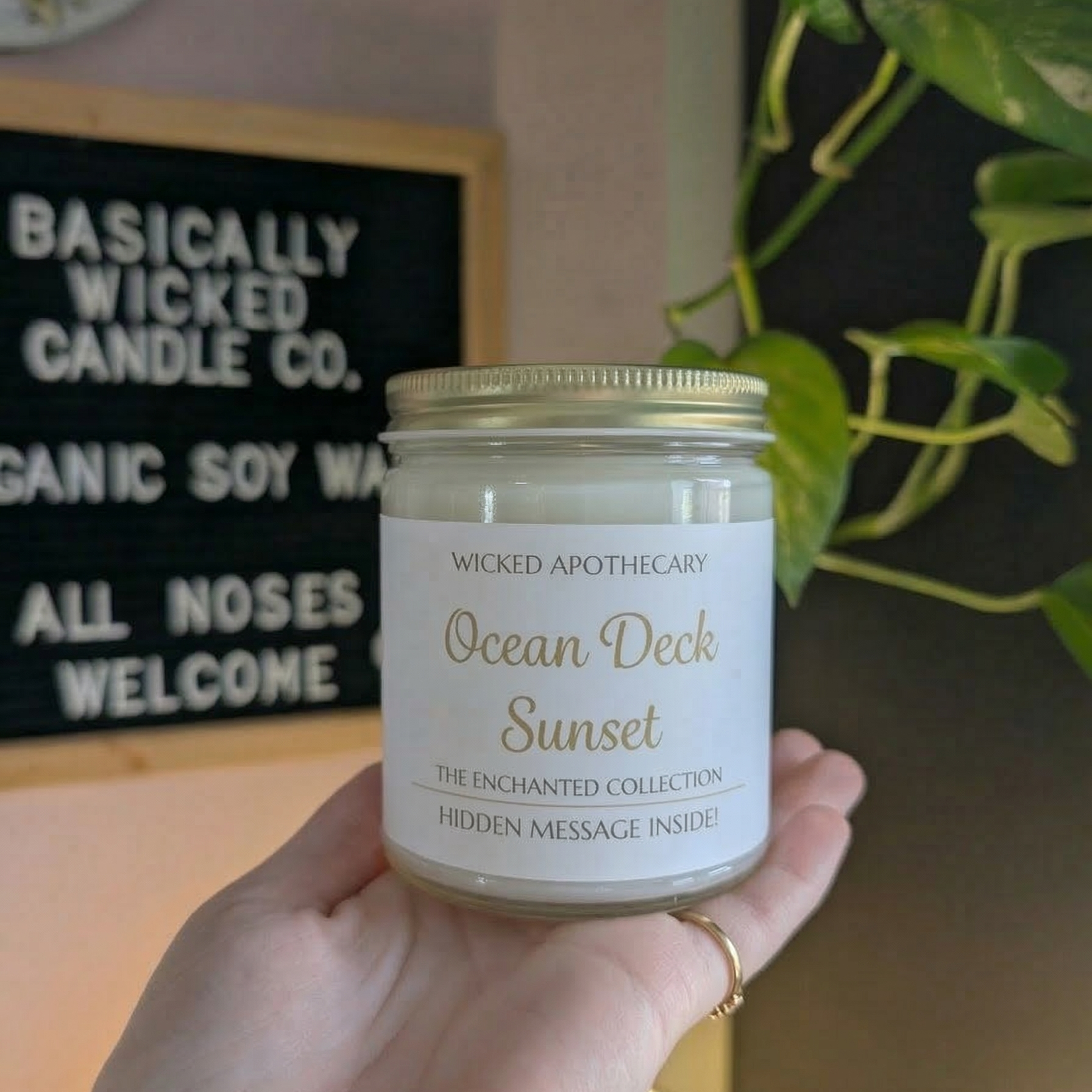 Hand holding a candle labeled 'Ocean Deck Sunset' with a blurred background featuring a sign and plant.
