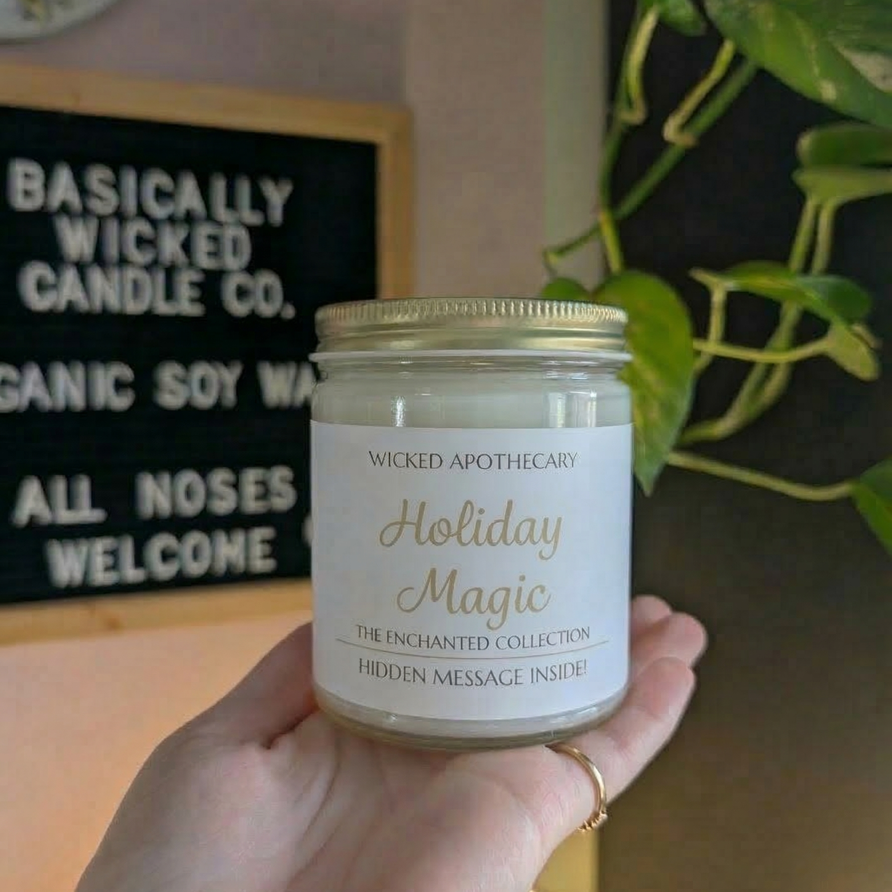 Hand holding a candle labeled 'Ocean Deck Sunset' with a blurred background featuring a sign and plant.