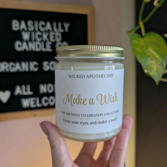 Hand holding a candle labeled 'Make a Wish' from Wicked Apothecary in front of a store sign.