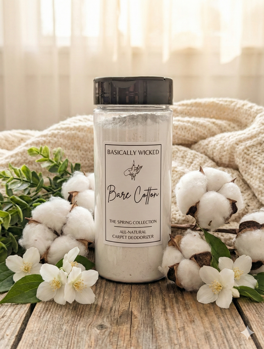 Bottle of 'Basically Wicked Bare Cotton' carpet deodorizer with cotton flowers and greenery on a wooden surface.