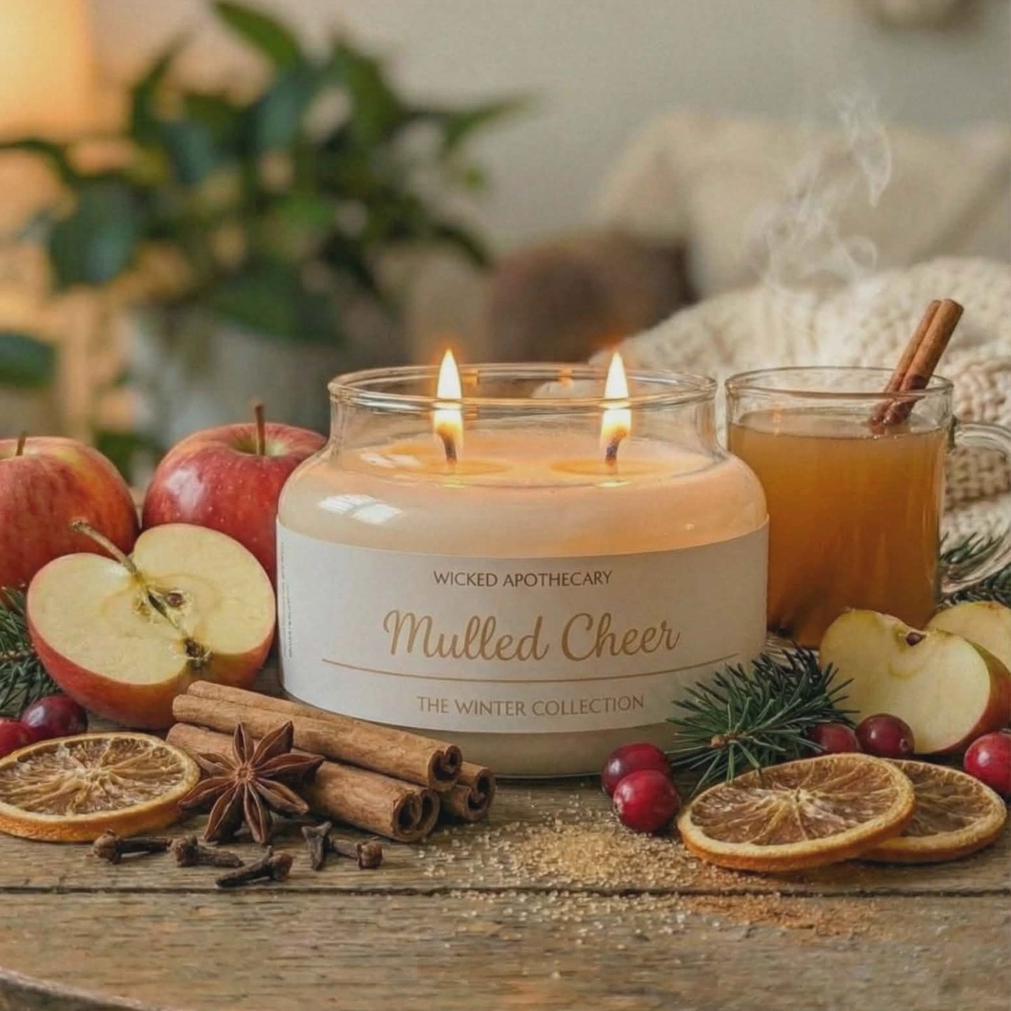 Candle labeled 'Mulled Cheer' on a table with apples, cinnamon, and oranges.