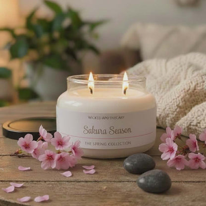 Candle in a jar labeled 'Sakura Season' on a wooden table with cherry blossoms and stones.