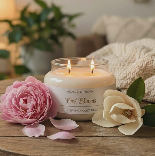 Candle labeled 'First Blooms' from Wicked Aromachery on a wooden surface with flowers.