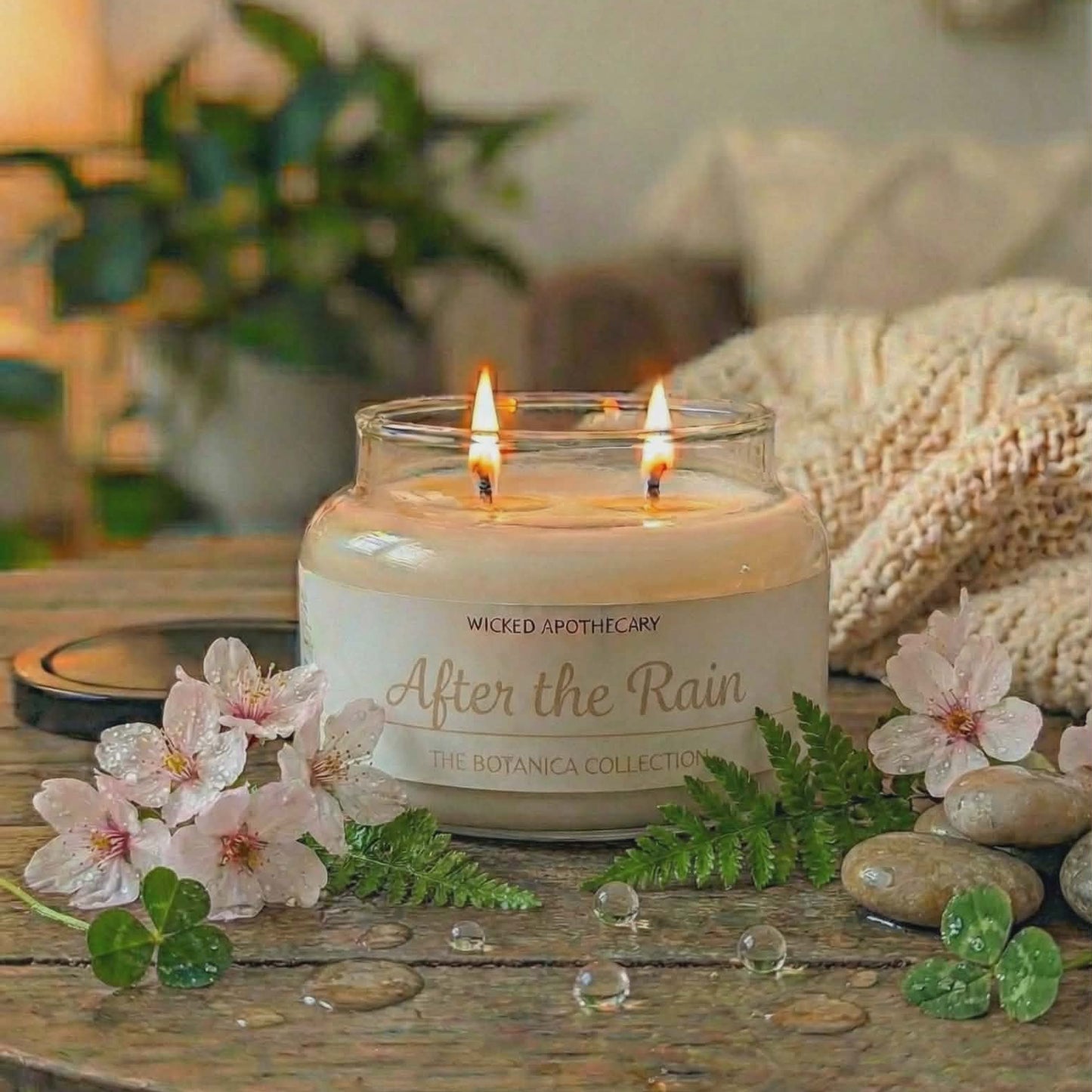 Candle labeled 'After the Rain' on a wooden table with flowers and pebbles.