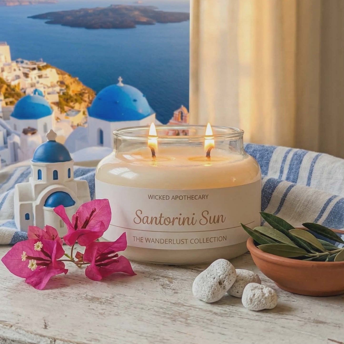 Santorini-themed candle on a table with flowers and stones, overlooking a scenic view.