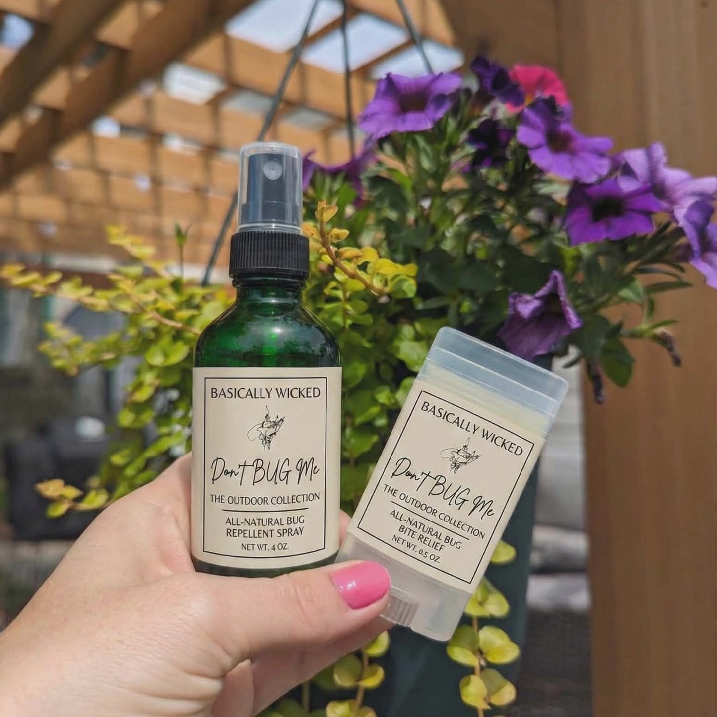 Hand holding a bottle and a small container of 'Basically Wicked' outdoor collection products with flowers in the background.