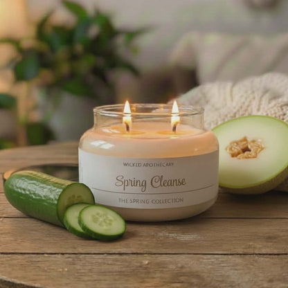 Candle labeled 'Spring Cleanse' on a wooden table with cucumbers and melon, blurred background
